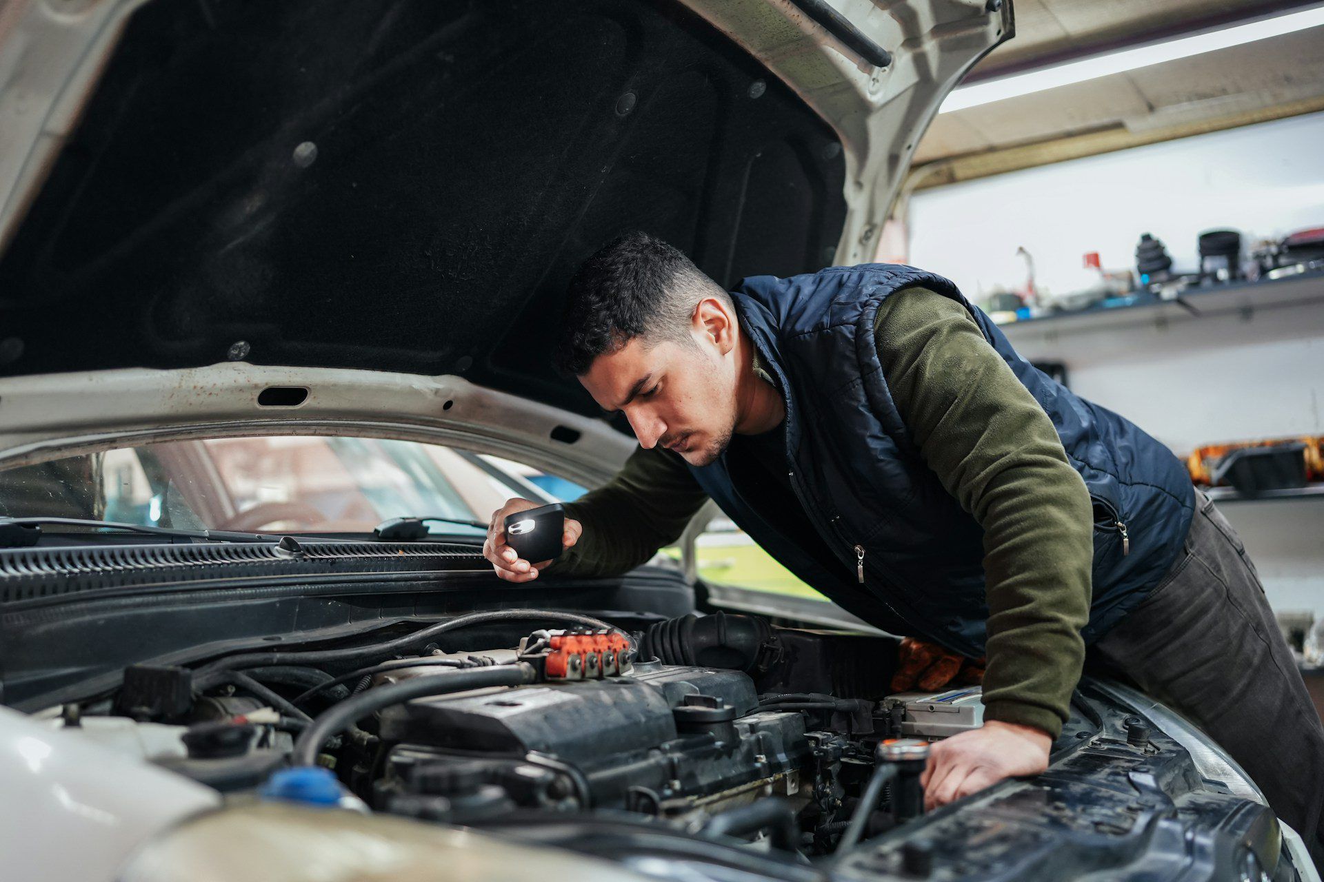 Man inspecting a car engine with a flashlight in a garage setting.