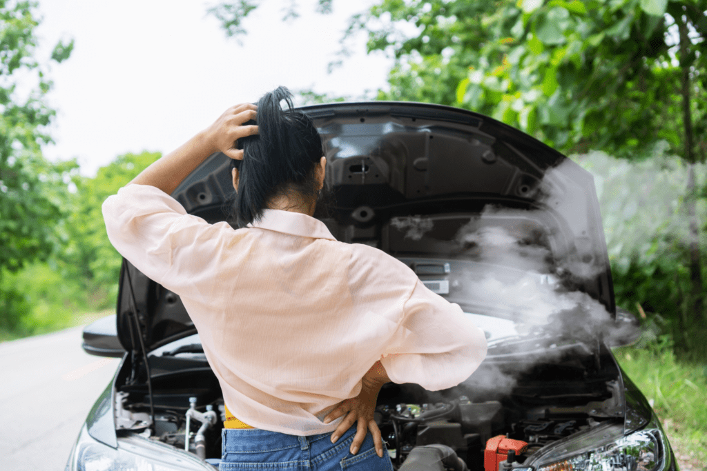 A woman stands facing her car with the hood up as smoke billows from underneath the hood because she needs engine repairs in Albuquerque. 