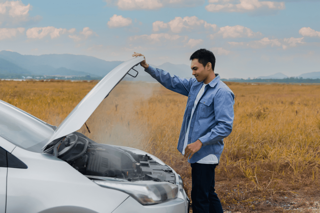 A man lifts his car hood to see smoke pouring out of the engine along the side of the road. 